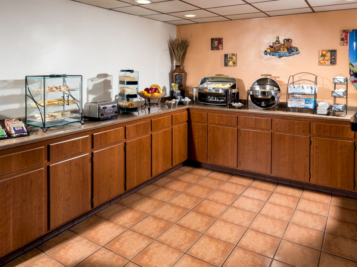 A breakfast buffet corner featuring a variety of food items and appliances, including a waffle maker, fruit, pastries, and beverages on countertops.