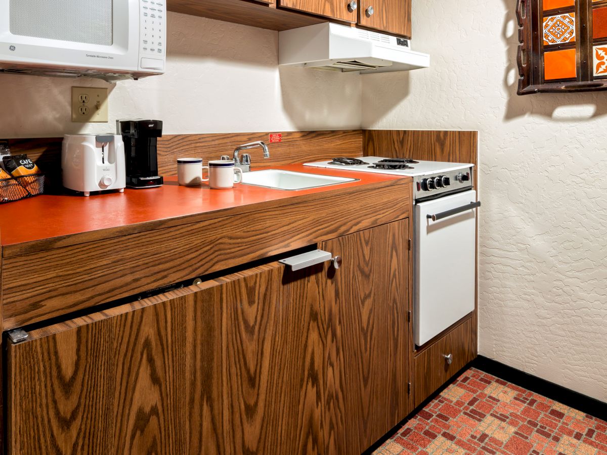 This image shows a small kitchen with wood cabinets, a microwave, a coffee maker, a stove, and a colorful tiled floor. The countertop is orange.