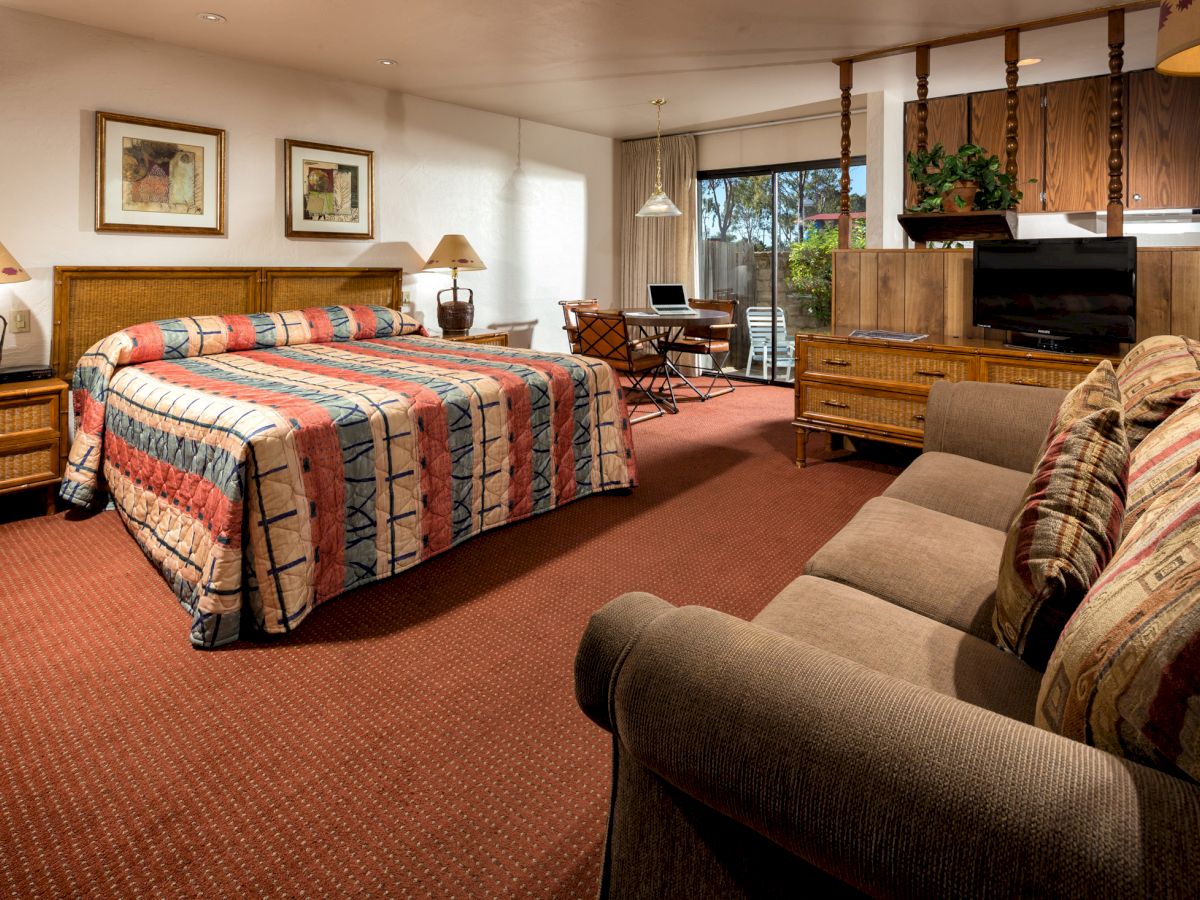 A cozy hotel room features a large bed with vibrant bedding, a sofa, a desk, a TV, and two framed artworks on the wall, with natural light.