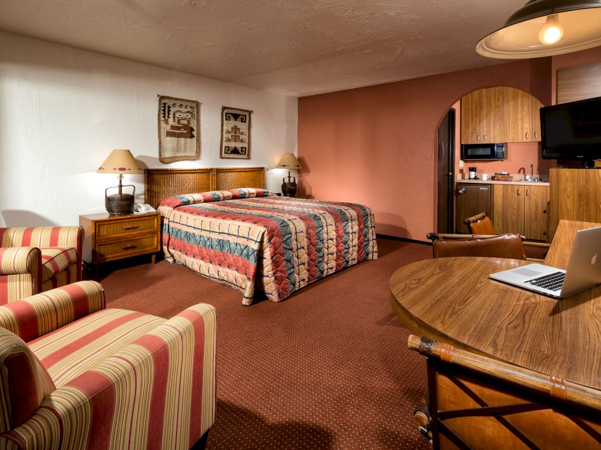 A cozy hotel room features a bed, two striped armchairs, a round table with a laptop, a kitchenette, and a wall-mounted TV.