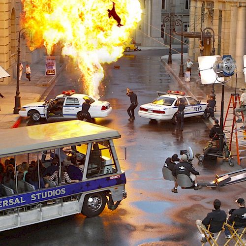 A Universal Studios tour bus observes a movie scene being filmed with police cars, cameras, lighting, and an explosion in the background.
