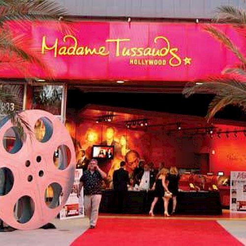 An entrance to Madame Tussauds Hollywood, featuring a red carpet, palm trees, large film reel decorations, and visitors.
