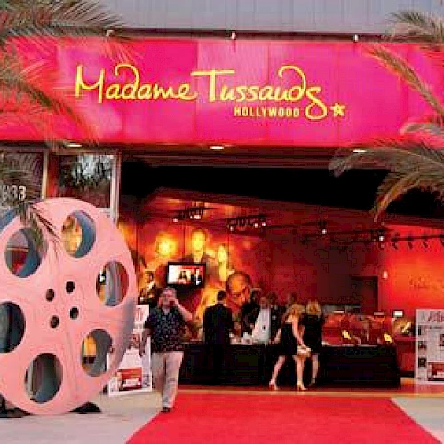An entrance to Madame Tussauds Hollywood, featuring a red carpet, palm trees, large film reel decorations, and visitors.