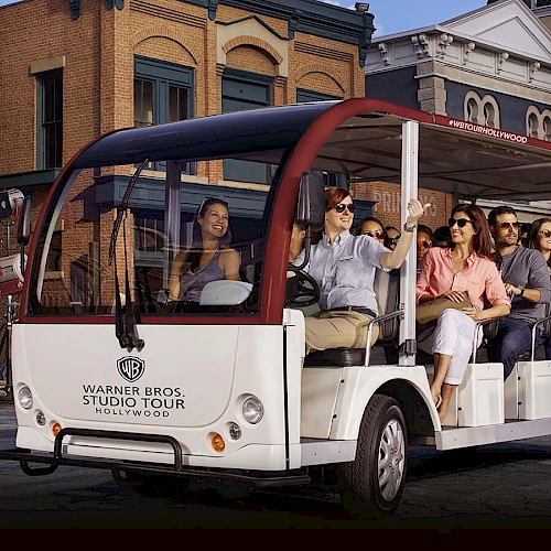 A group of people sits in an open-air tram labeled "Warner Bros. Studio Tour" while touring a set designed like a city street with various buildings.