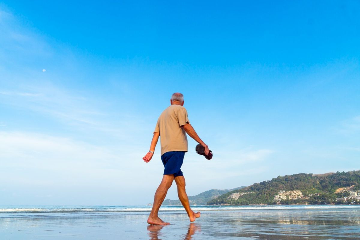 An older man is walking barefoot along a beach, holding his shoes, with the ocean and hills in the background under a clear blue sky.