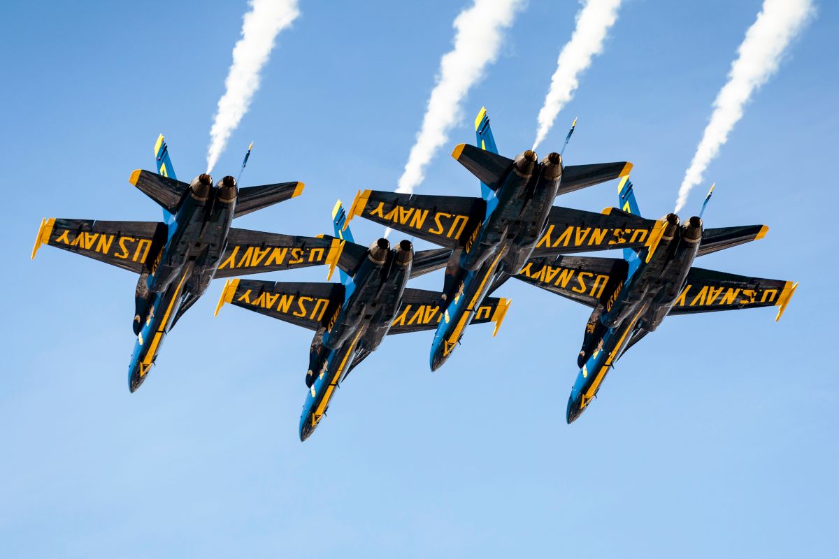 Four U.S. Navy jets perform an aerial formation with contrails visible against a clear blue sky, showcasing precision flying.