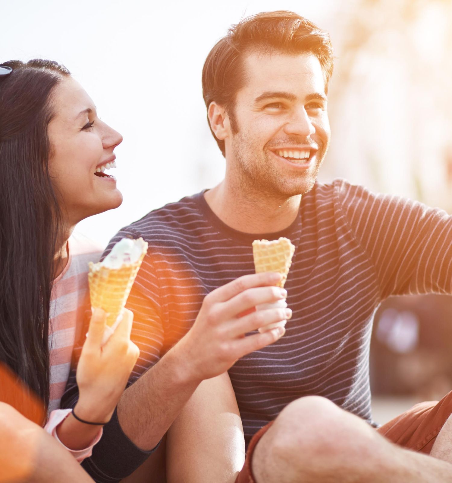 A man and woman are sitting outdoors, enjoying ice cream cones, and smiling in the sunlight. They appear to be happy and relaxed.