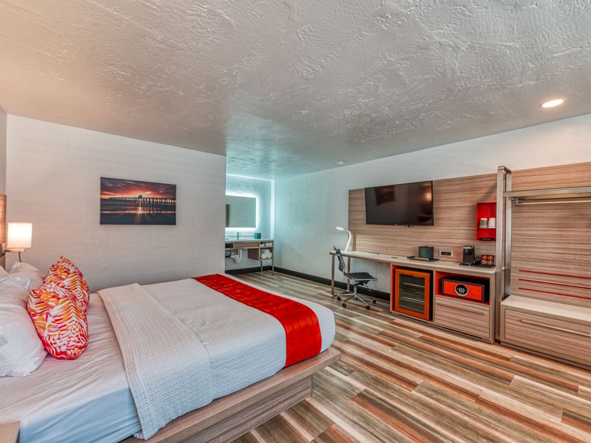 A modern hotel room with a bed, vibrant pillows, a wall-mounted TV, desk, and wardrobe, featuring stylish wood flooring and decor.