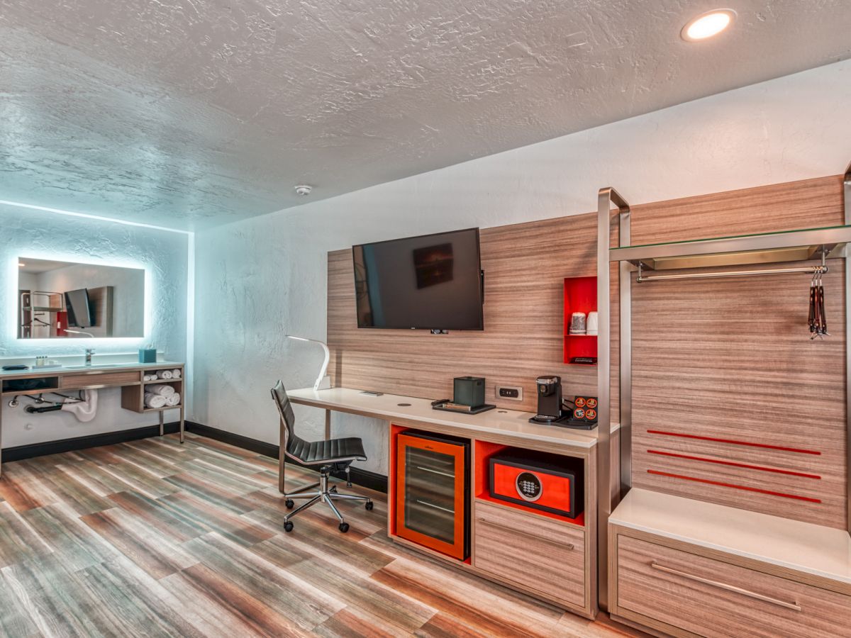 Modern hotel room with a wall-mounted TV, desk, chair, coffee maker, mini refrigerator, open wardrobe, and illuminated mirror.