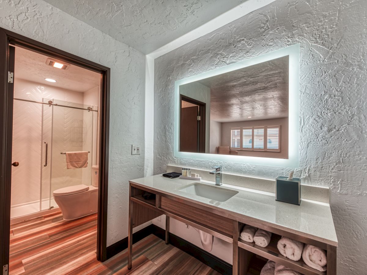 A modern bathroom with a large illuminated mirror, sink, and storage underneath; visible shower and toilet in an adjacent room on the left.