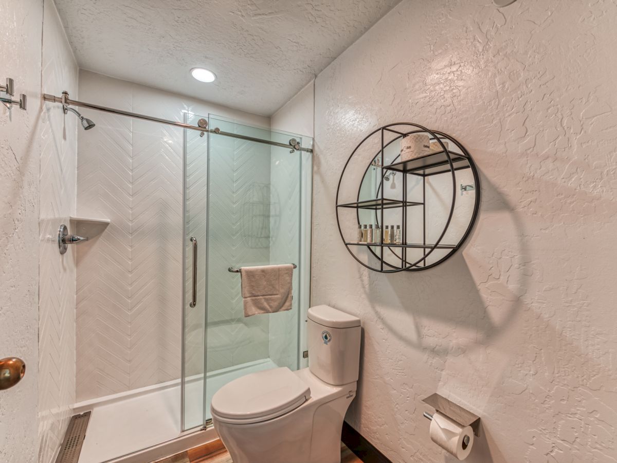 A bathroom with a glass-enclosed shower, toilet, round wall shelf, and toilet paper holder on a white textured wall, seen with wooden accents.