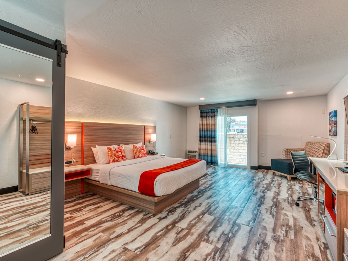 A modern hotel room featuring a large bed with red accents, a seating area, a desk, and a large window with a view.