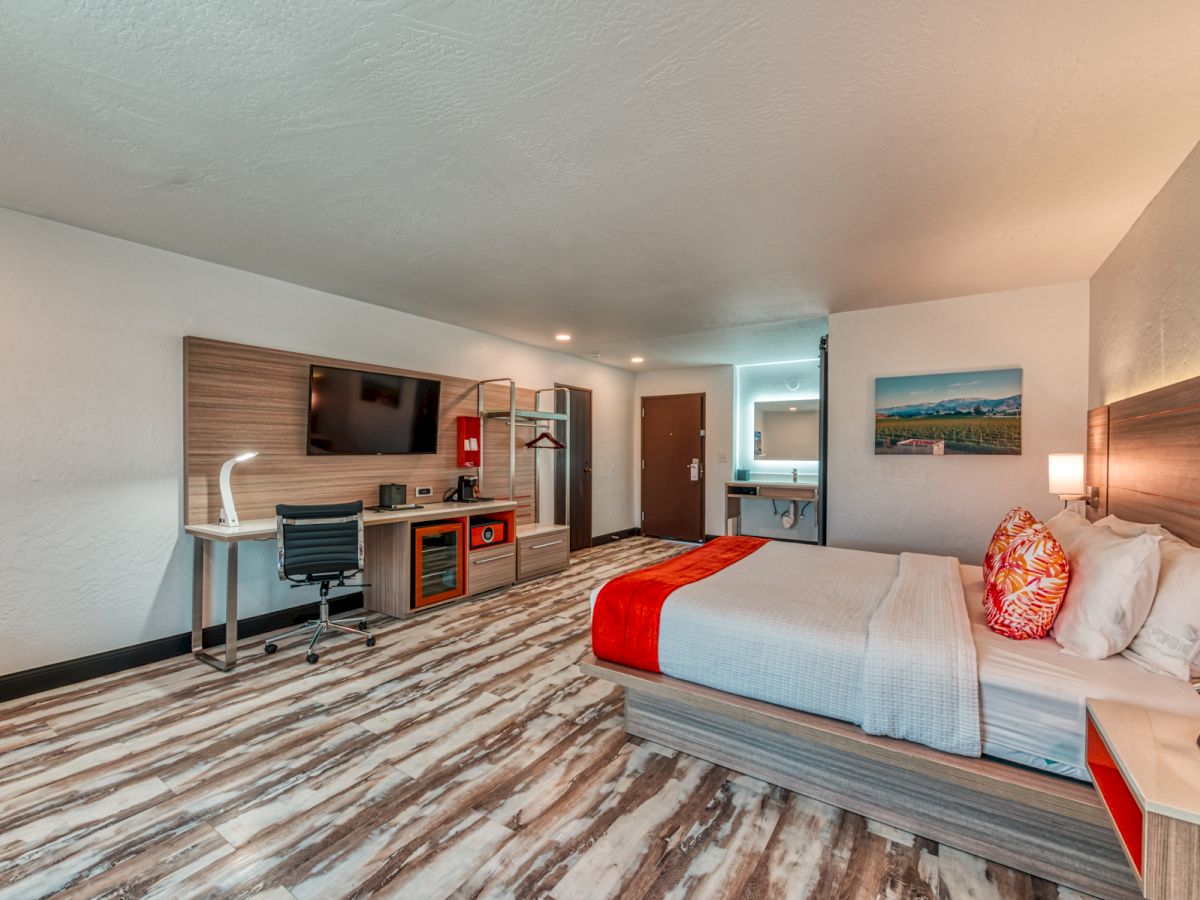 The image shows a modern hotel room with a bed, desk, TV, and a bathroom area. The decor is contemporary, with wood accents and bright colors.