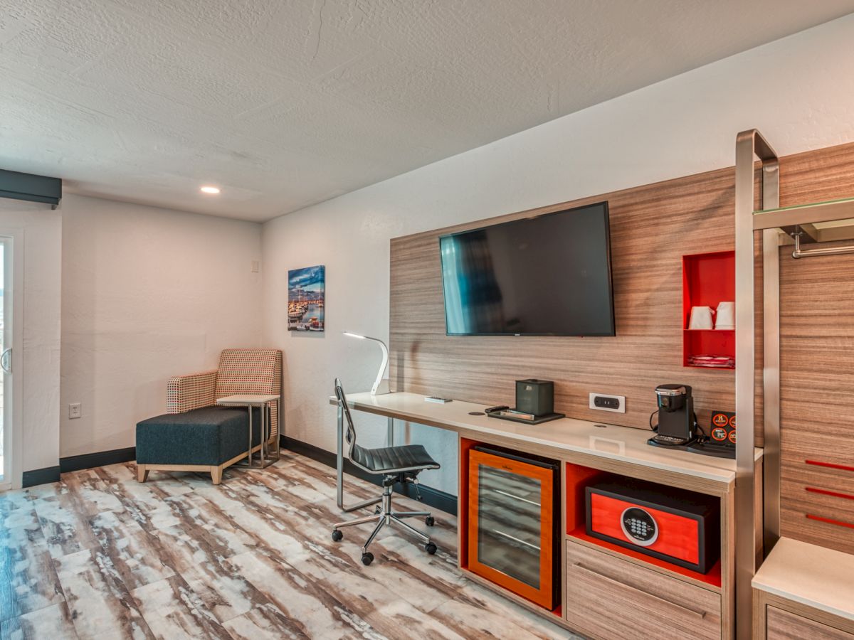 This image shows a modern hotel room with a desk, chair, TV, lounge chair, coffee maker, and a small refrigerator.