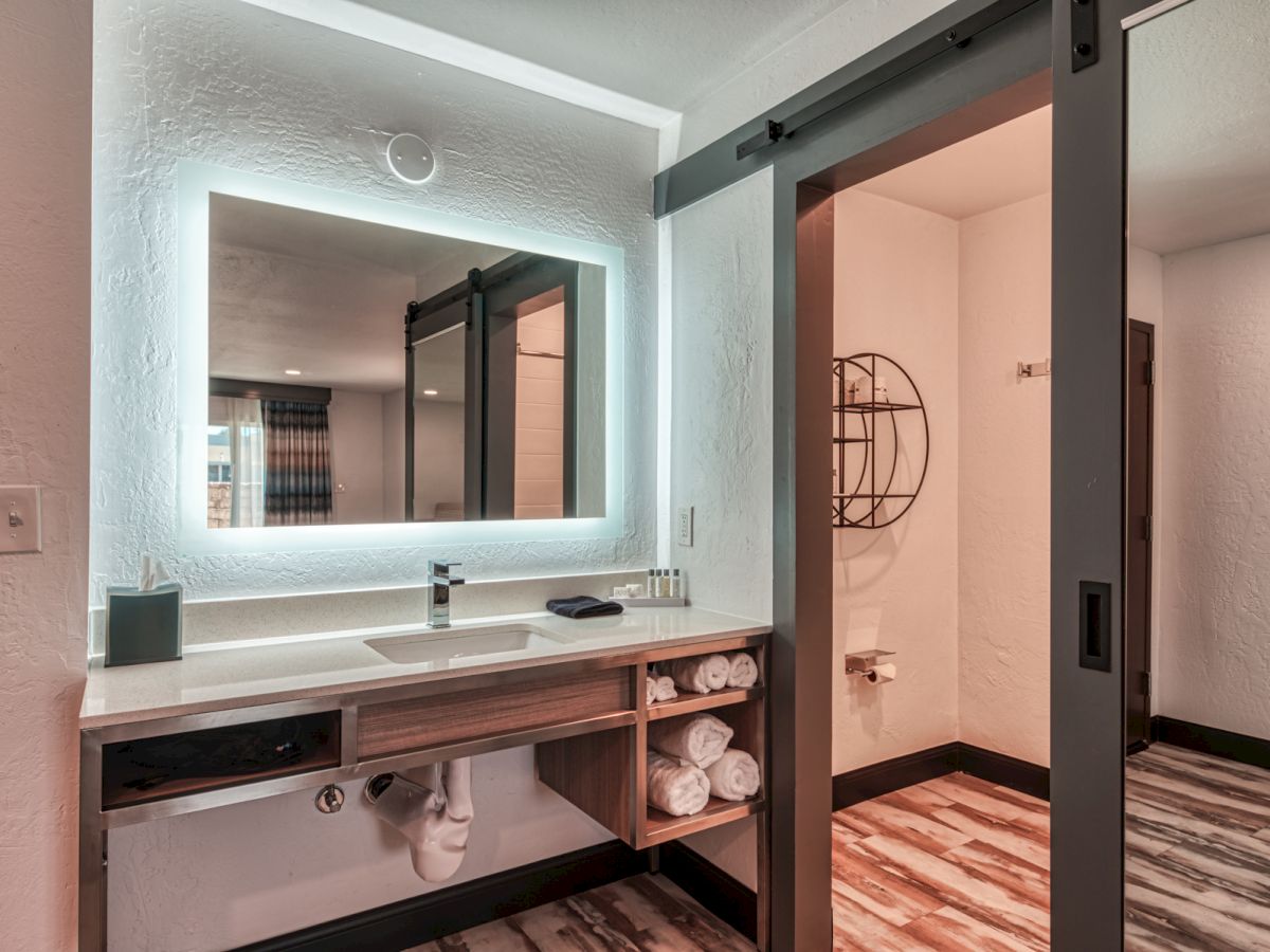 A modern bathroom features a backlit mirror above a sink with towels underneath, next to a sliding door leading to the toilet area.