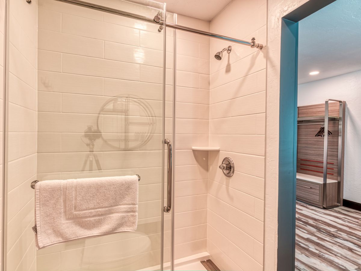 This image shows a modern bathroom with a glass shower door and white tiles, featuring a white towel hanging on the door. The adjacent room is also visible.