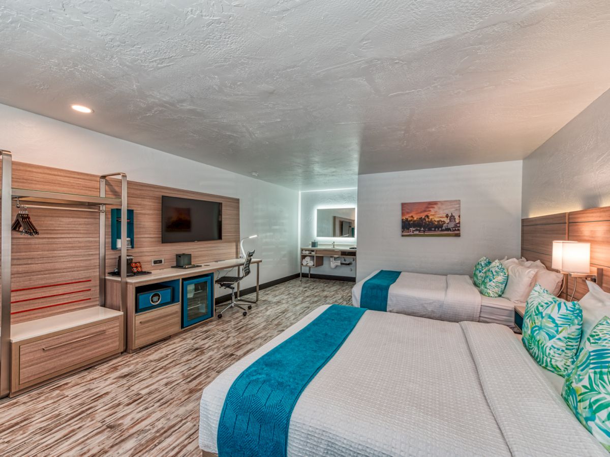 A modern hotel room with two queen beds, a TV, a desk, and a kitchenette area. The room is brightly lit with a contemporary design ending the sentence.