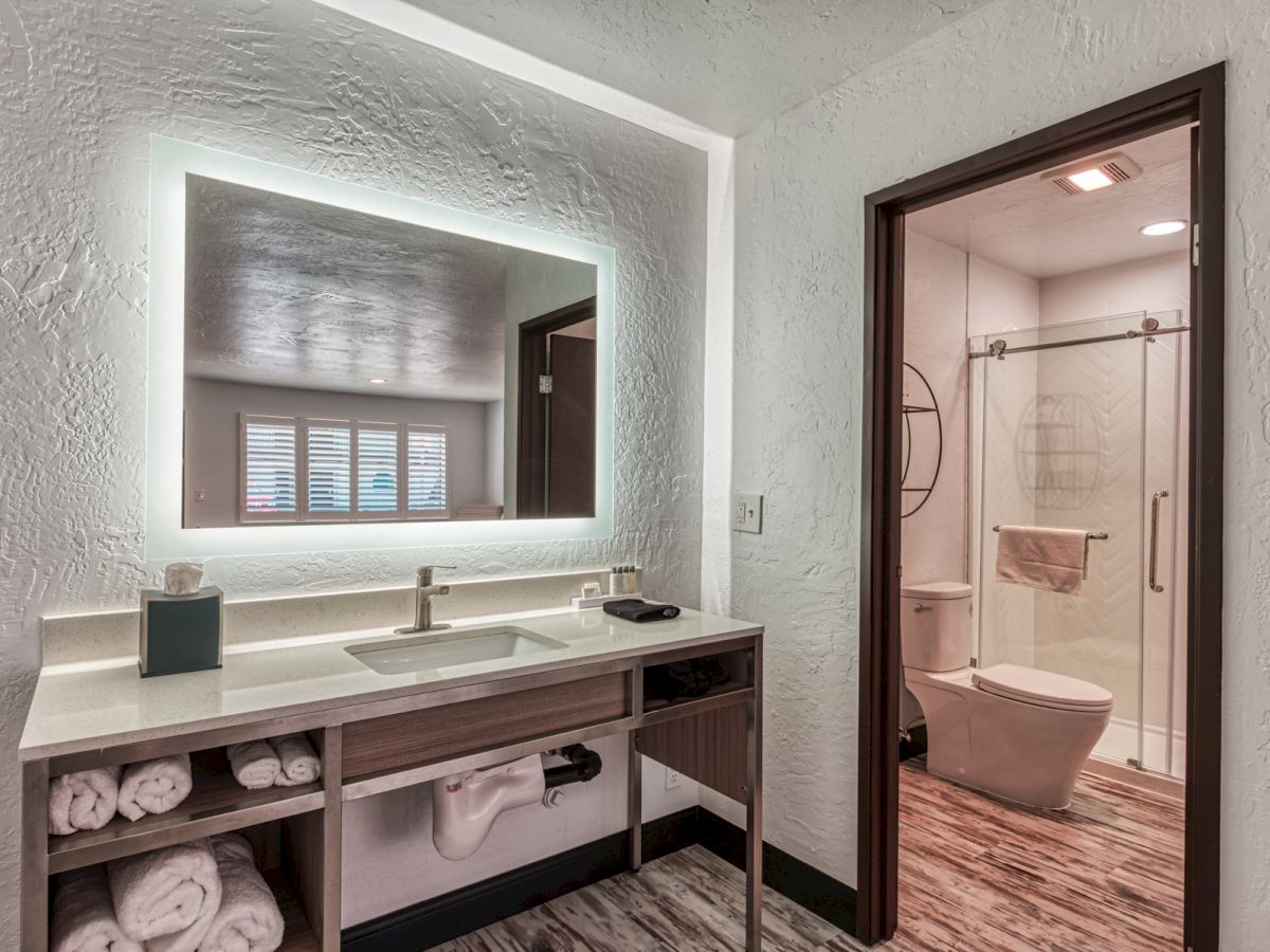 Modern bathroom with a lit mirror, sink, neatly stacked towels, and toiletries on a counter, leading to a shower and toilet area with glass doors.