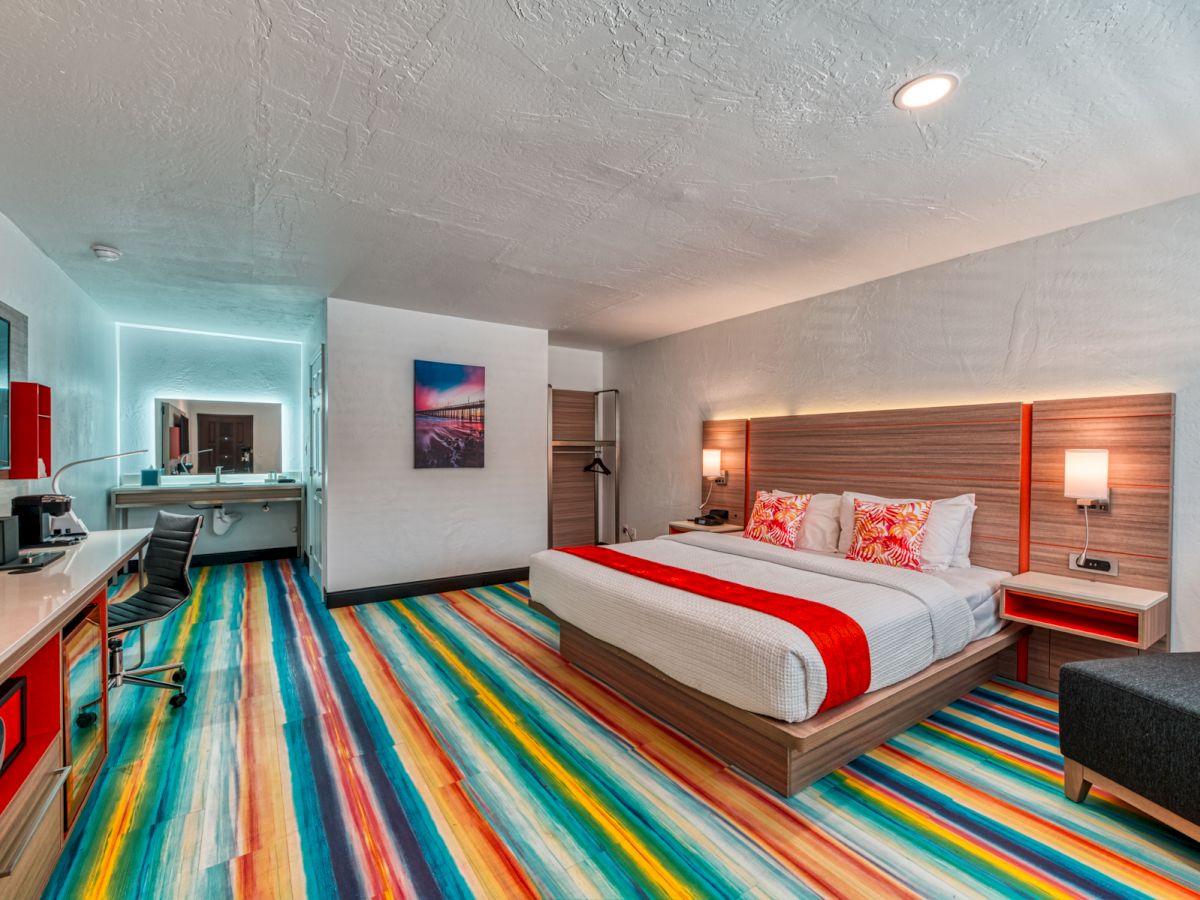 A modern hotel room with a king-size bed, colorful striped carpet, work area, and a bathroom in the background.