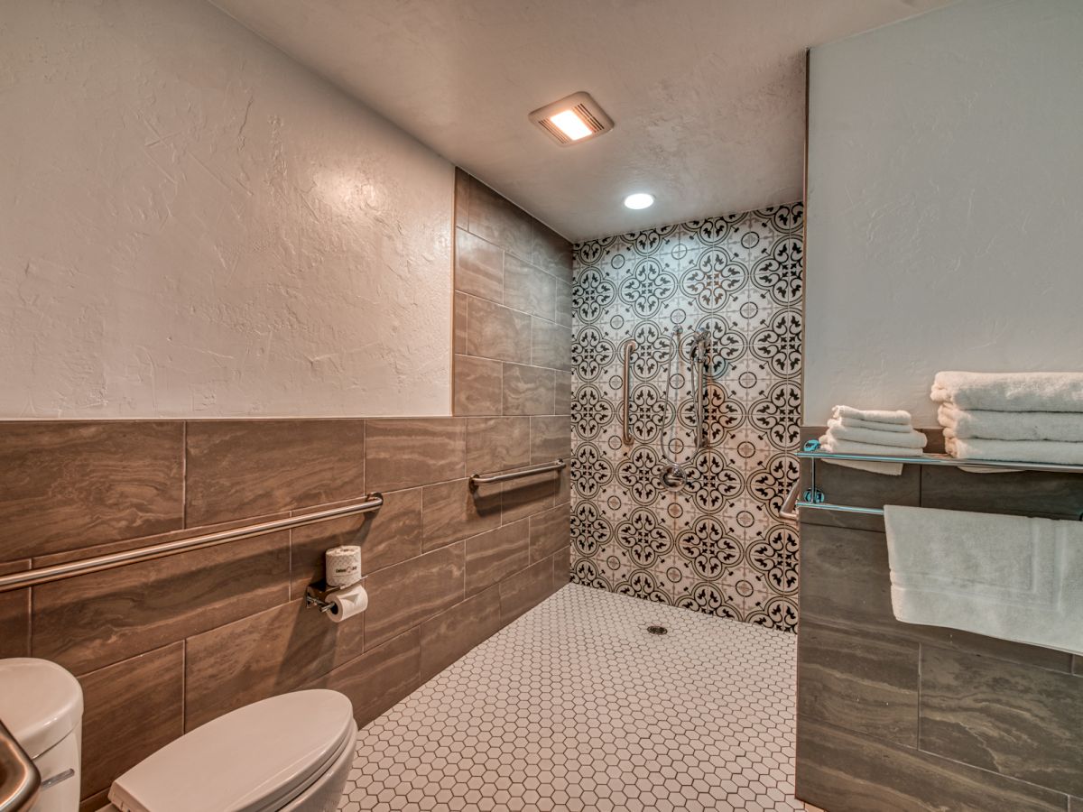 A modern bathroom features a toilet, a roll of toilet paper, a walk-in shower with patterned tiles, and a towel rack with folded towels.