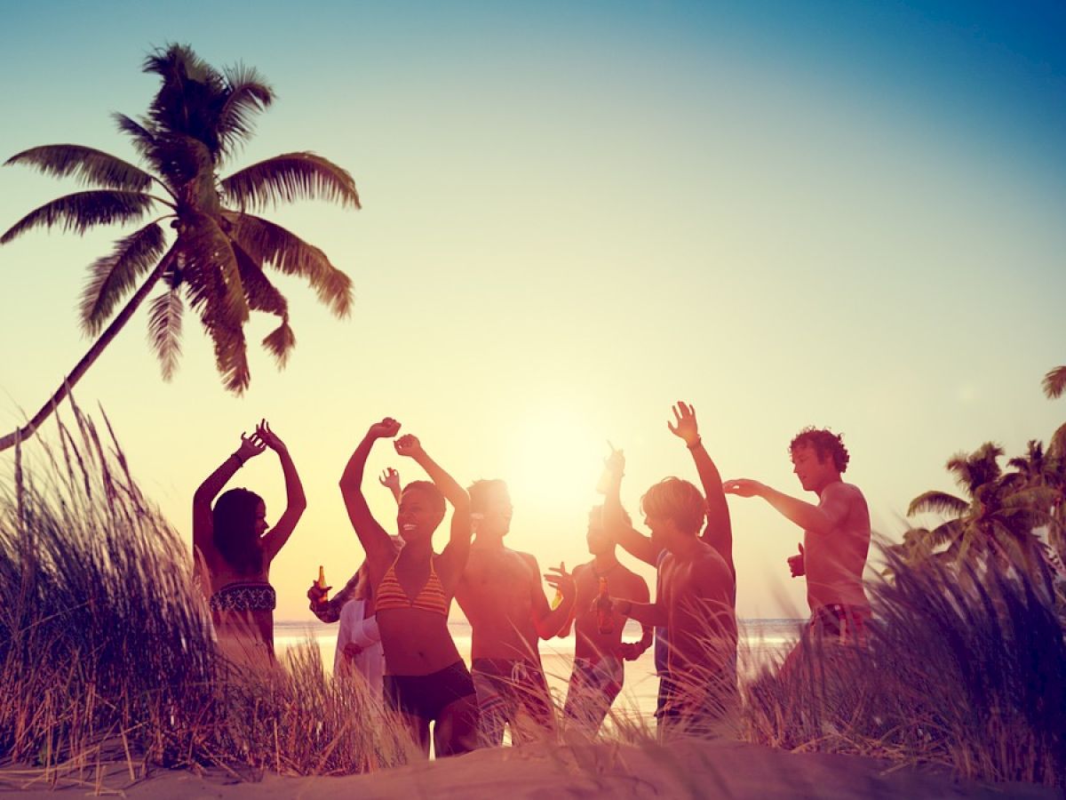 A group of people celebrating at the beach during sunset, with palm trees and sand dunes in the background.