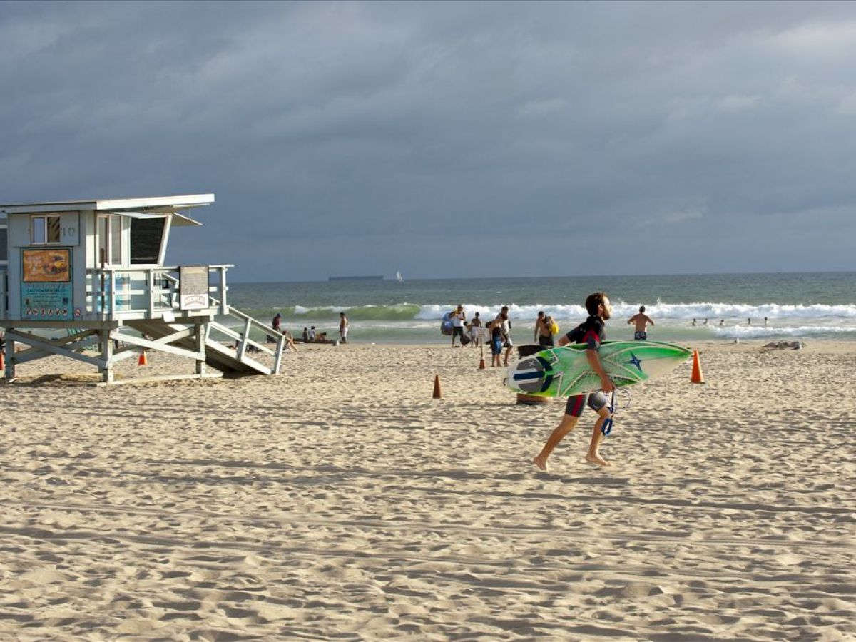 A lifeguard tower, beachgoers, and a surfer with a board are on a sandy beach with the ocean in the background, under a cloudy sky.