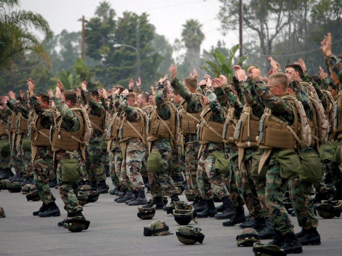 A large group of people in military uniforms performs a synchronized exercise outdoors, with helmets on the ground in front of them.