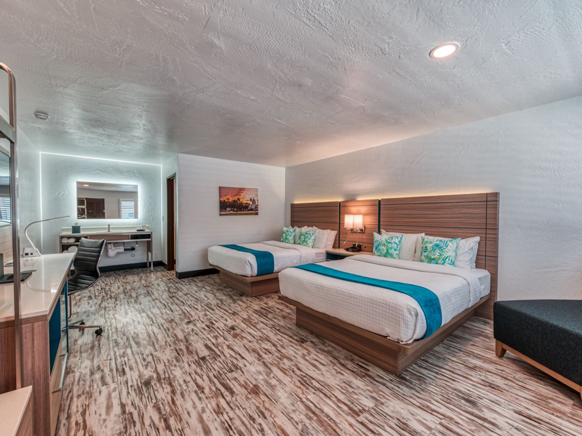 This image shows a clean, modern hotel room with two beds, a desk, a large mirror, and a seating area. The room has wood flooring and contemporary decor.