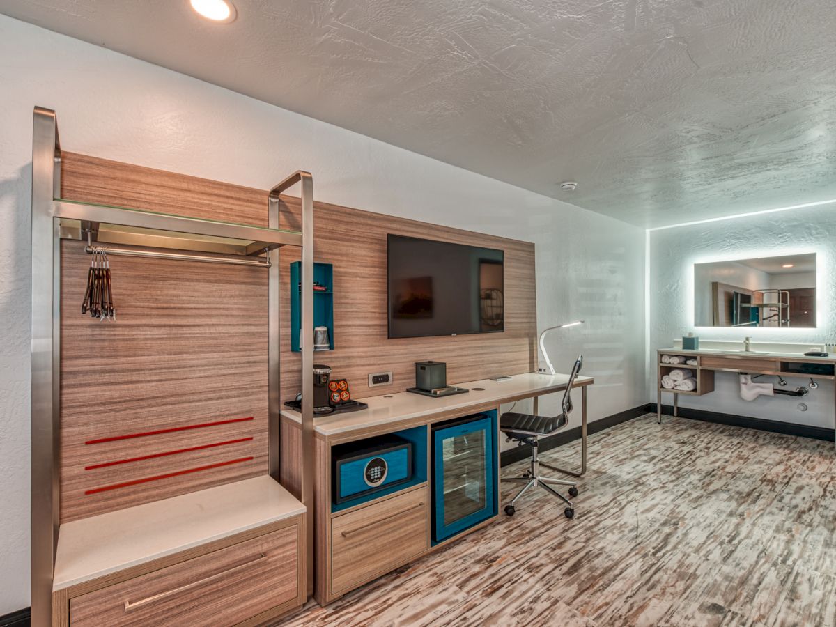 A modern hotel room with a work desk, office chair, TV, mirror, coat rack, mini-fridge, and shelves, featuring contemporary design elements.