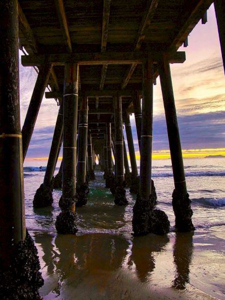 A stunning image of a pier taken from underneath at sunset, showcasing colorful skies, reflective wet sand, and waves gently hitting the shore.