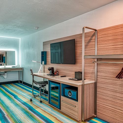 The image shows a modern hotel room with a desk, chair, TV, open wardrobe with hangers, colorful striped flooring, and a mirror with lighting.