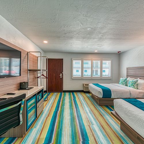 A modern hotel room with two beds, a desk, a large TV, a coffee maker, and vibrant striped flooring. The room has a cozy, bright, and clean ambiance.