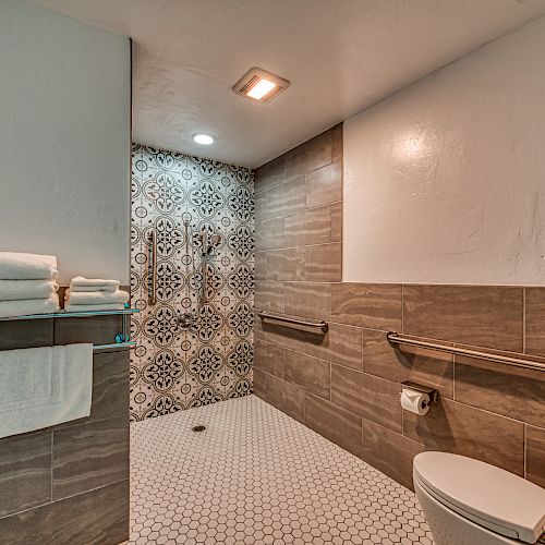 A modern bathroom with a walk-in shower, patterned tiles, towel rack, and handrails for accessibility. The overall design is sleek and functional.