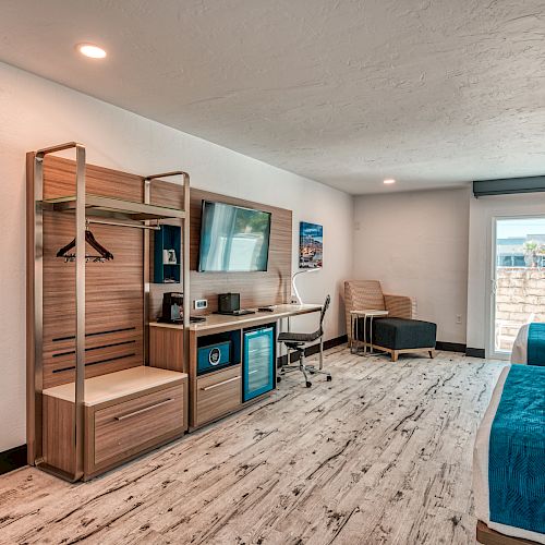 A modern hotel room with two beds, a TV, a desk, a mini-fridge, and an open wardrobe space. Natural light enters through a window.