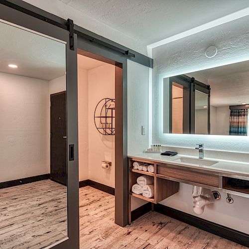 The image shows a modern bathroom with a sliding door, illuminated mirror, sink with storage space, towels, and toiletries neatly arranged.