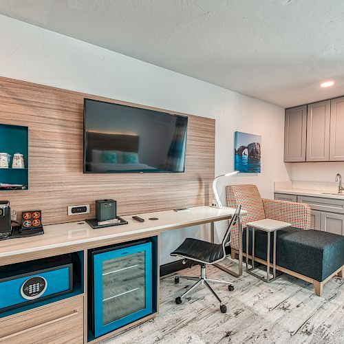 A modern room features a wall-mounted TV, desk with chair, shelving, coffee machine, microwave, sink, and storage cabinets.