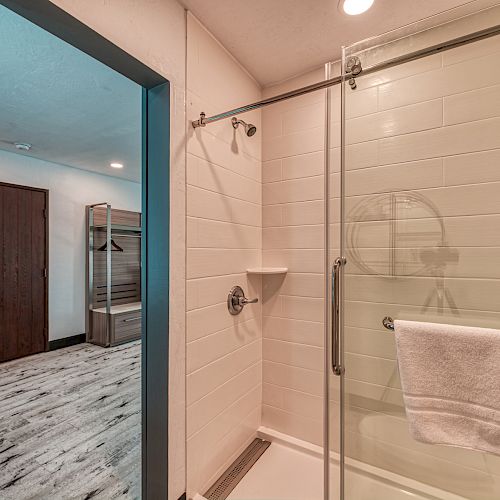 The image shows a bathroom with a shower enclosure featuring a glass door and white tiles, leading to a room with wooden flooring and furniture.