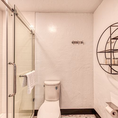 The image shows a clean, modern bathroom with a glass-enclosed shower, a toilet, and a circular metal shelf with toiletries on the right wall.