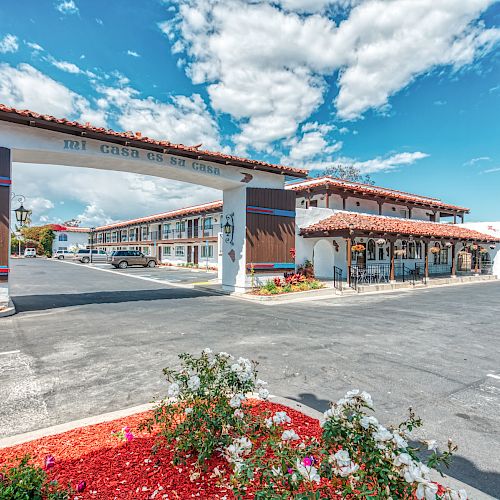 The image shows an entrance to a motel with cars parked, a manicured flower bed, and clear skies, creating a welcoming atmosphere.