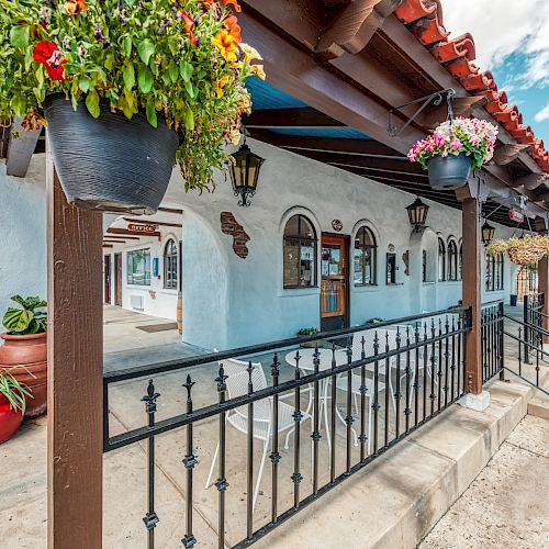 The image shows an exterior view of a charming building with white walls, a red-tiled roof, hanging flower pots, and decorative iron railings.