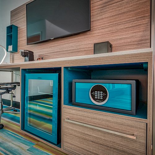 A modern hotel room desk setup includes a chair, mini-fridge, safe, lamp, TV, and colorful flooring.