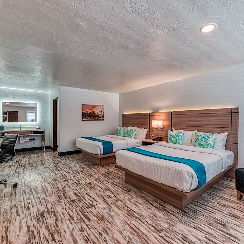 This image shows a hotel room with two beds, a desk, a chair, a TV, and a mirror. The decor includes blue accents and modern furniture.