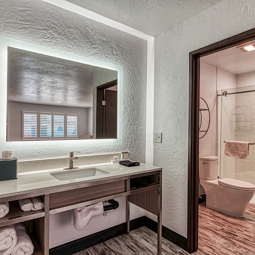 A modern bathroom with a backlit mirror, sink, and towels on a shelf; a toilet and shower are visible through an open doorway ending the sentence.