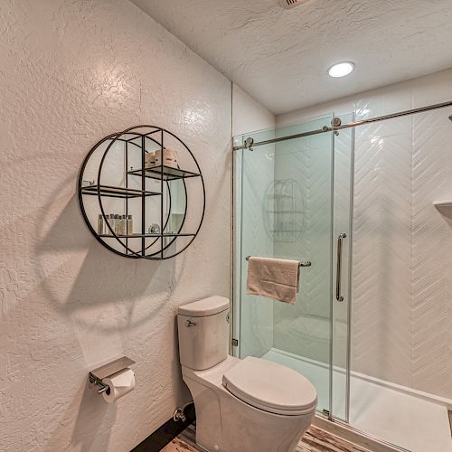 This image shows a modern bathroom with a white toilet, a glass-enclosed shower, a circular wall shelf, and a towel hanging on a rack.