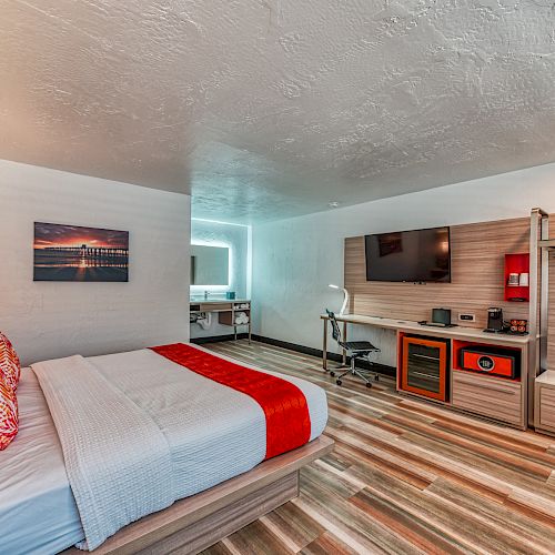 A modern hotel room featuring a bed, a wall-mounted TV, a desk, a minibar, and a wardrobe with an open design, in a warm, neutral color palette.