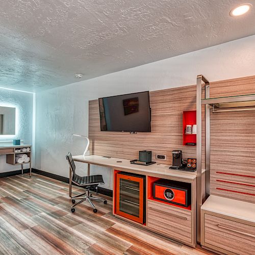 A modern hotel room features a desk, TV, mini-fridge, chair, and clothing storage, with a well-lit vanity area in the back.