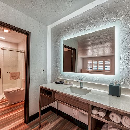 This image shows a modern bathroom with a large illuminated mirror, sink, and storage. The shower area with a glass door is in the background.