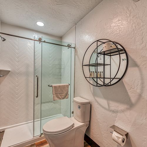 A modern bathroom with a glass-enclosed shower, toilet, circular shelf, and wall-mounted toilet paper holder on stucco walls, is depicted in this image.