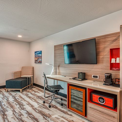 The image shows a modern hotel room with a TV, desk, chair, coffee machine, and a small fridge. There is also a seating area next to the window.