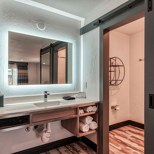 A modern bathroom with a large backlit mirror, wooden countertop with sink, toiletries, and towels. A sliding door leads to a toilet area.